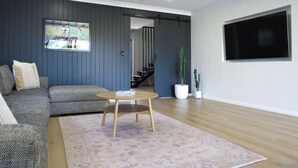 Un salon moderne avec un canapé gris, une table basse ronde en bois, un ventilateur de plafond, un téléviseur à écran plat et un escalier menant à un autre niveau.