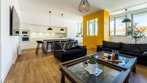 Ett modernt vardagsrum med en svart lädersoffa, ett glasbord med en växt och en köksdel med vita skåp och en gul accentvägg.