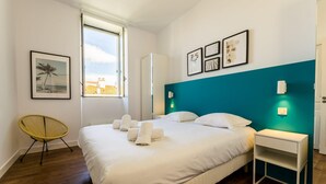 Ett hotellrum med en säng, en stol, ett sängbord och ett fönster med utsikt över byggnader.