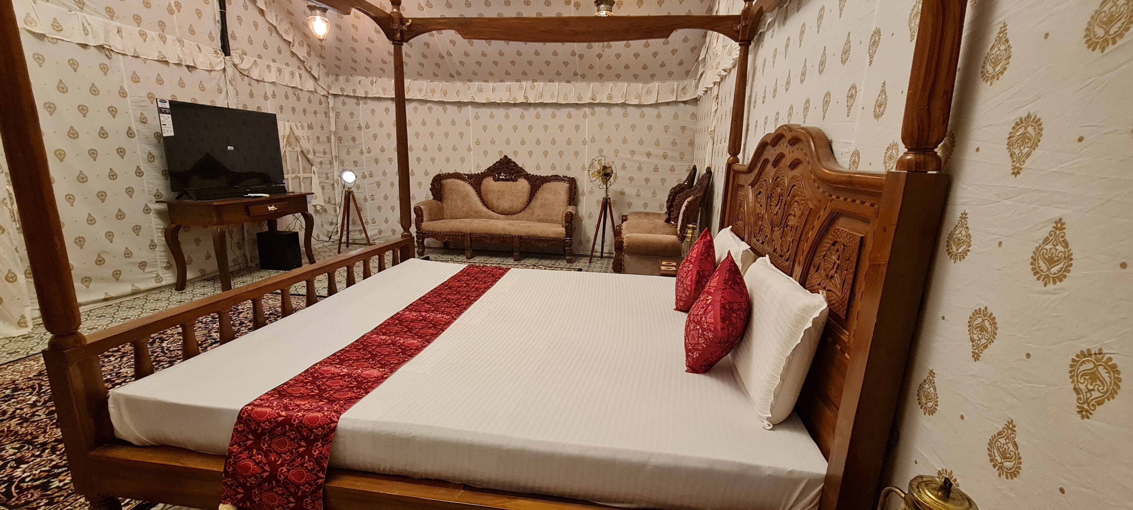 Una cama con dosel y soporte de cuatro postes, con cabecera de madera, una manta roja y almohadas rojas. A la izquierda, una tocador con espejo y lámpara. En el fondo, un sofá contra la pared.