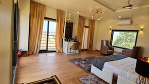 A spacious bedroom with a large bed, a desk, and a chair. There is a window with a view of trees and a ceiling fan. - Stay Serene Luxury  (Murbad)