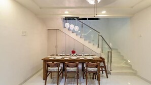 A dining area with a large wooden table set for six, a staircase with glass railings, and a modern chandelier. - Stay Serene Luxury  (Murbad)