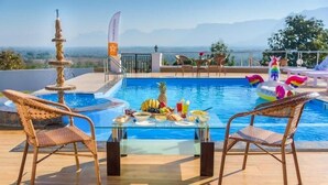 A poolside area with wicker chairs, a table with fruits and drinks, and a view of mountains. - Stay Serene Luxury  (Murbad)