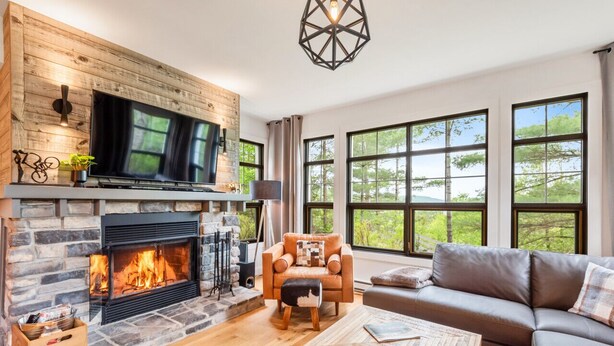 A cozy living room with a stone fireplace, a comfortable sofa, and large windows offering a view of the outdoors. - Tremblant Prestige - Les Eaux 235-1 (Mont-Tremblant)