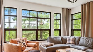 A modern living room with large windows, a leather sofa, a leather armchair, and a wooden coffee table. - Tremblant Prestige - Les Eaux 235-1 (Mont-Tremblant)