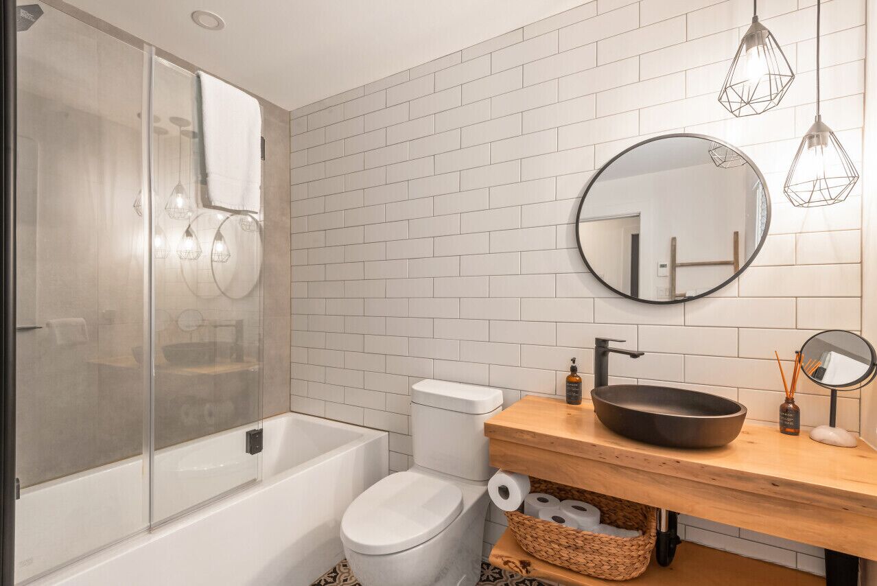 Un bagno moderno con cabina doccia in vetro, un water bianco, un mobile bagno in legno con specchio rotondo e un lavandino nero.