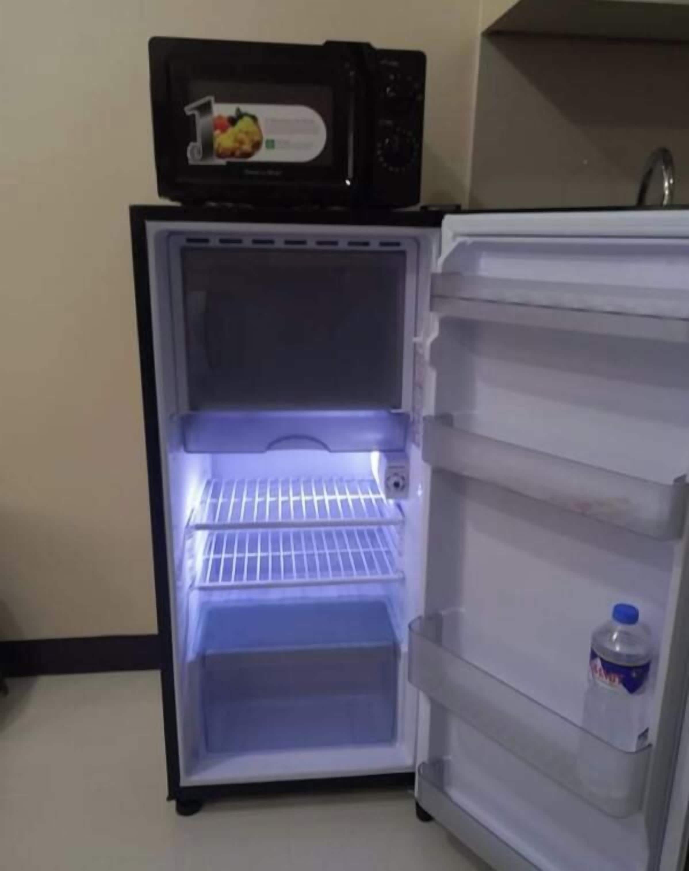 An open refrigerator with a microwave on top, a water bottle inside, and a shelf with a fruit basket.