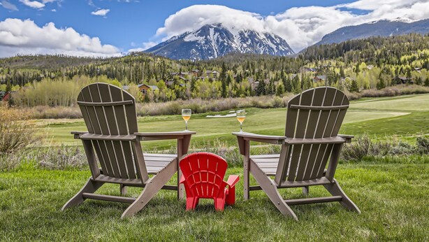 Two Adirondack chairs with a table and two glasses of wine, set on a grassy area with a mountain view.