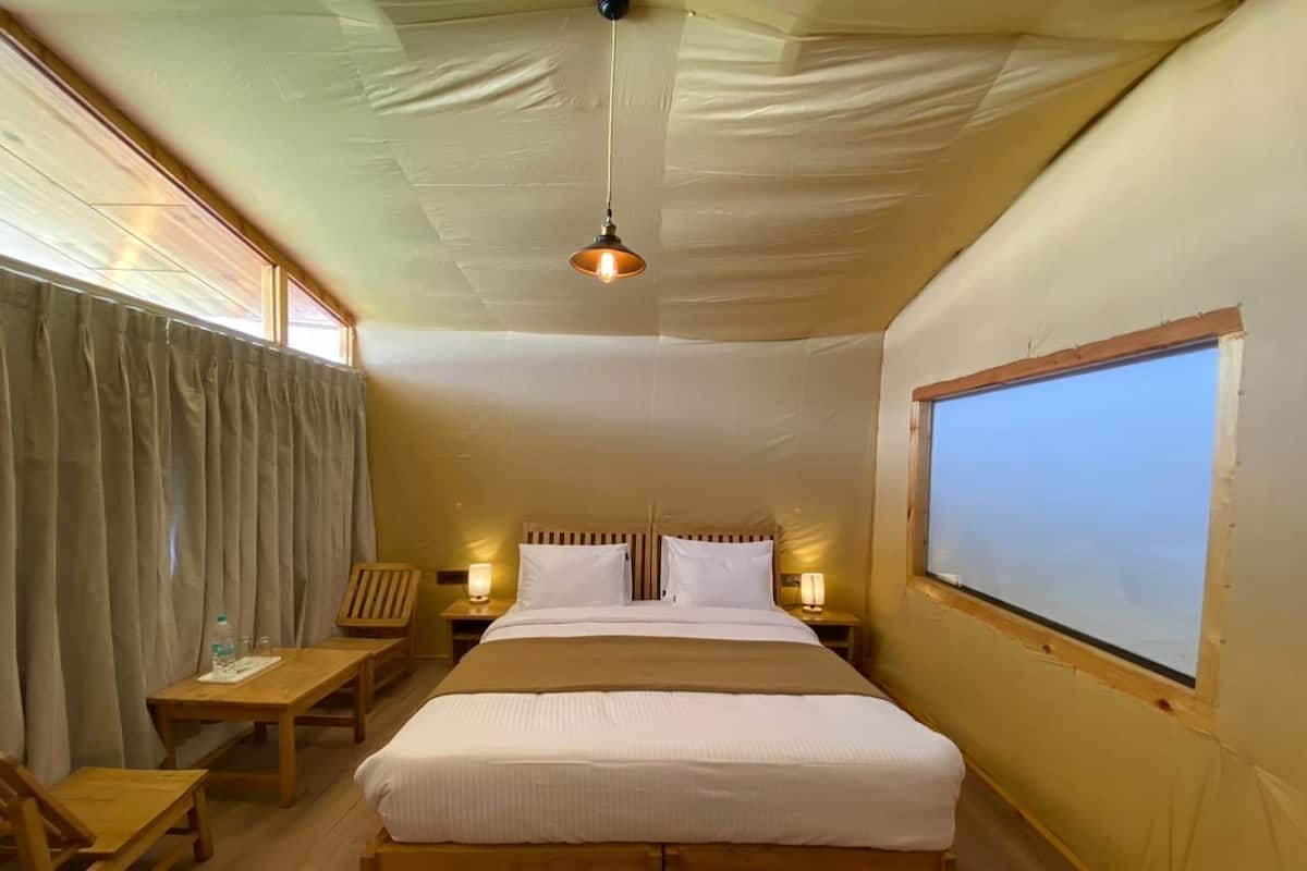 Habitación de hotel con una cama grande, cabecero de madera, dos mesitas de noche con lámparas, un escritorio de madera y un ventanal con cortinas.