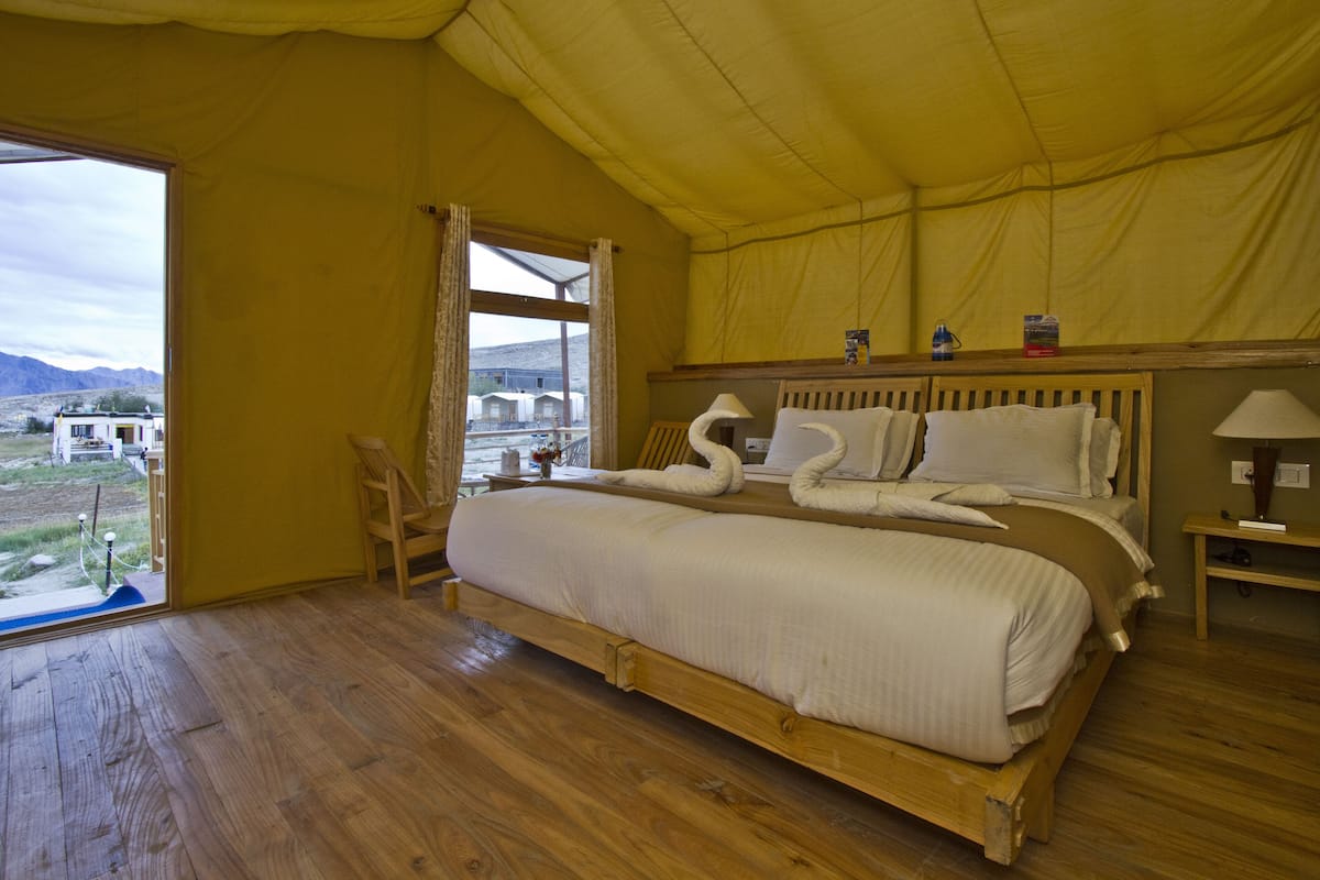 Una cama grande con estructura de madera y ropa de cama blanca, una mesita de noche de madera con una lámpara, y una ventana con cortinas.