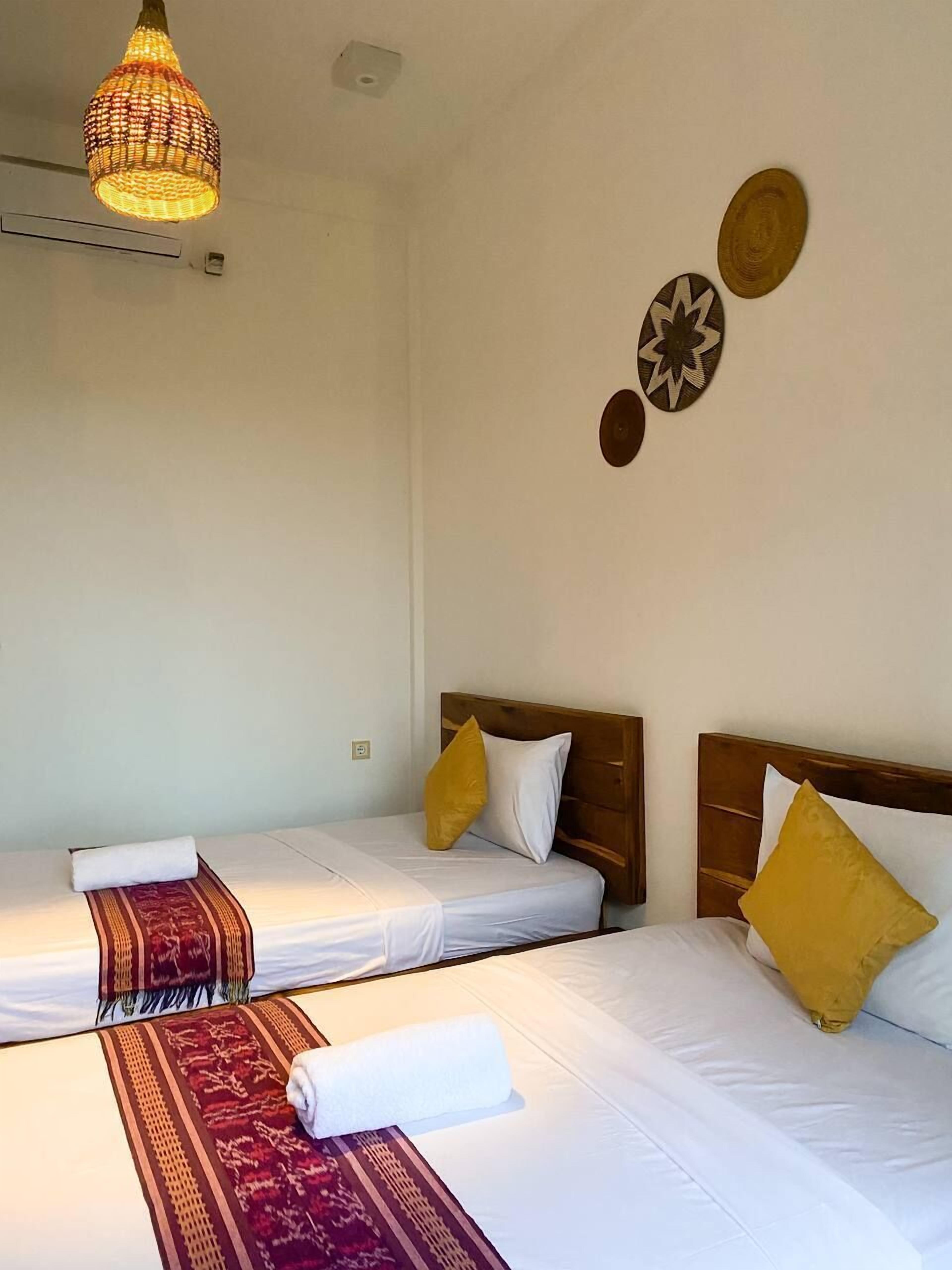 Habitación de hotel con dos camas individuales, cada una con cabecero de madera, ropa de cama blanca y una manta decorativa estampada. Hay una lámpara colgante sobre las camas.