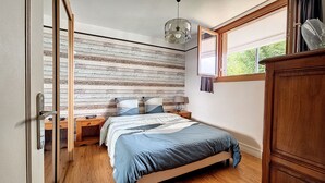 A bedroom with a wooden floor, a bed with a blue and white bedspread, a wooden headboard, a nightstand with a lamp, and a window with a view of greenery. - Appartement Centre-ville Proche Paris et Versailles (Sèvres)