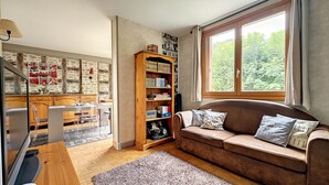 A cozy living room with a brown sofa, a wooden bookshelf, and a large window with a view of greenery. - Appartement Centre-ville Proche Paris et Versailles (Sèvres)