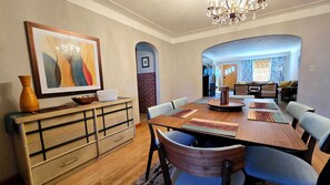 Un comedor con una mesa de madera, sillas y una araña. Hay un florero y un tazón sobre la mesa, y un cuadro en la pared.