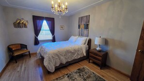 Un dormitorio con una cama, una silla, una mesita de noche con lámpara, una araña, un ventanal con cortinas y un cuadro en la pared.