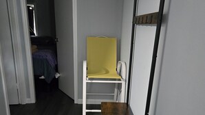 Um quarto com piso de madeira, uma cadeira branca, uma pequena mesa, uma tela amarela e uma estante de metal na parede.