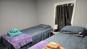 Duas camas de solteiro com roupa de cama cinzenta, um cobertor roxo e uma pilha de toalhas laranja.