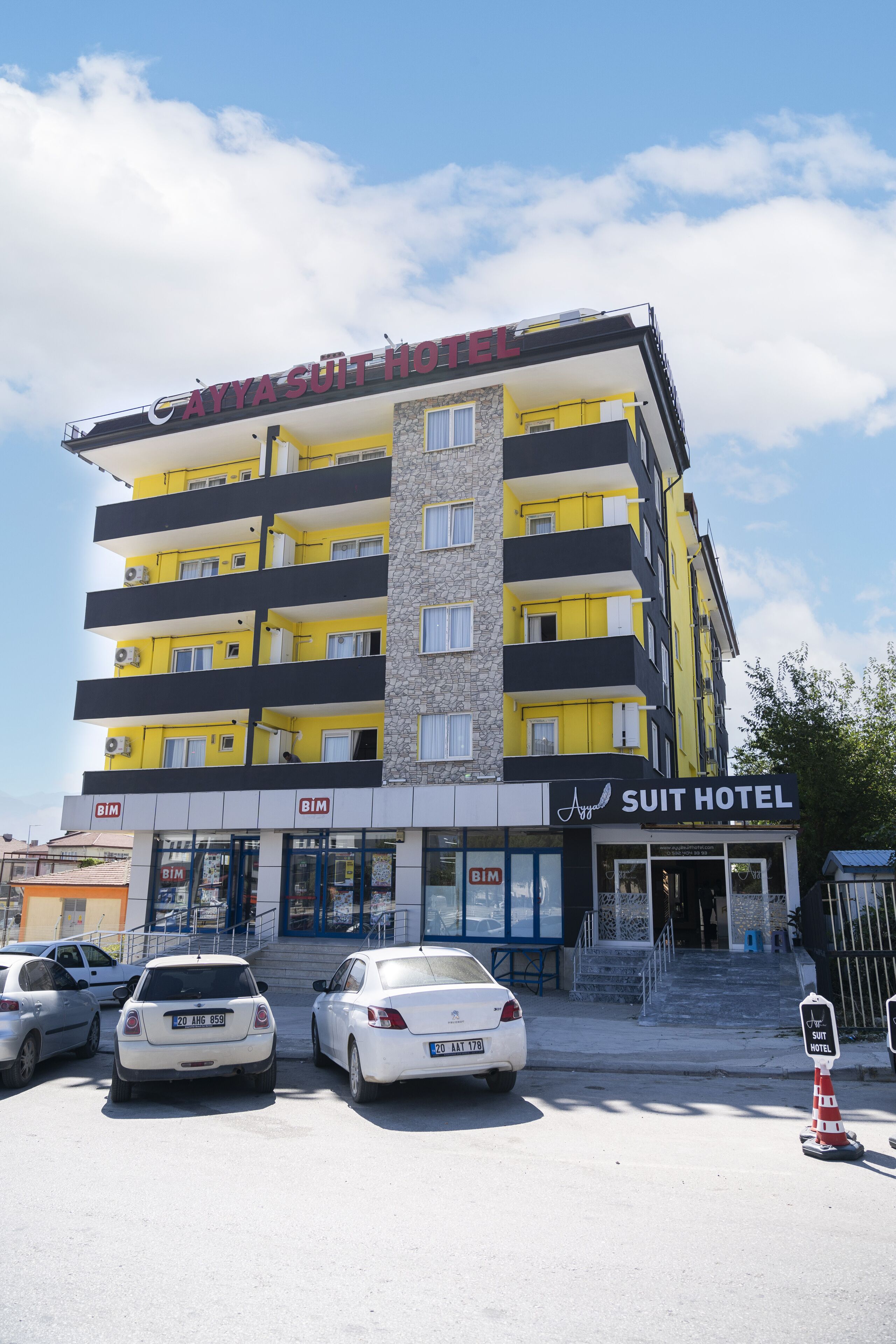 Un edificio de varios pisos con un cartel que dice “Suit Hotel”. Hay dos coches estacionados frente al edificio, el cual tiene varios balcones.