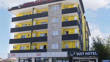 Un edificio de varios pisos con un cartel que dice “Suit Hotel”. Hay dos coches estacionados frente al edificio, el cual tiene varios balcones.