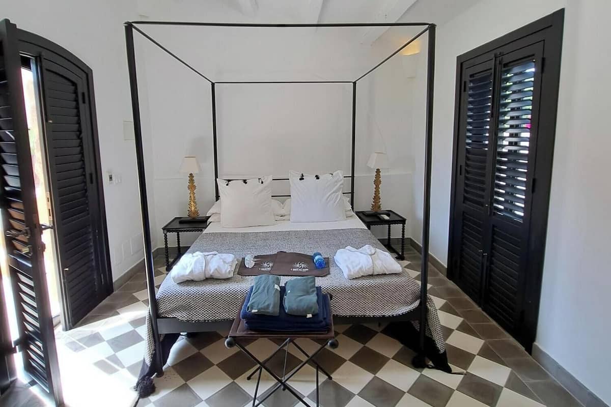 A four-poster bed with a checkered floor, two bedside tables, and a small table with towels and a bottle.