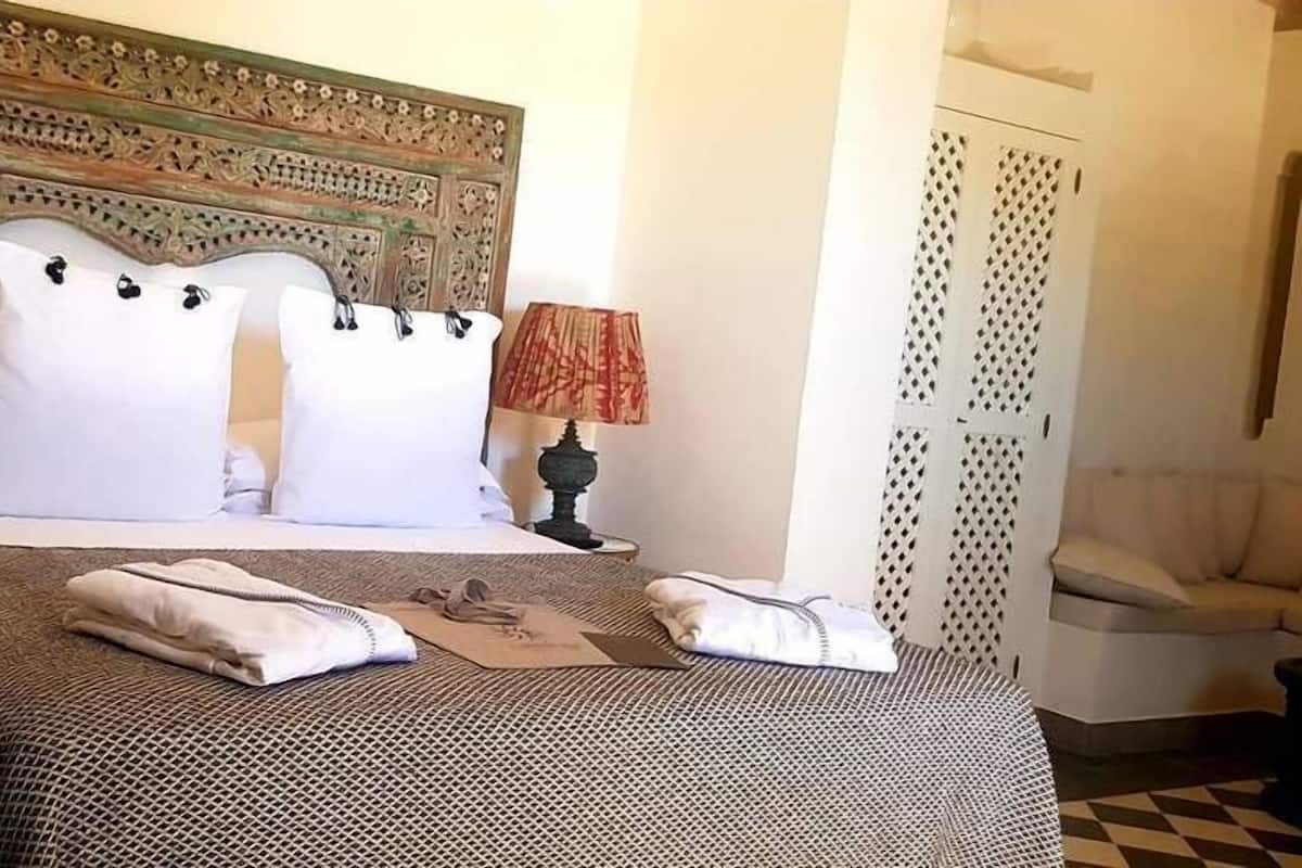 A neatly made bed with a patterned bedspread, two white pillows, and a folded towel on the bed. There are two lamps with red and white lampshades on either side of the bed.
