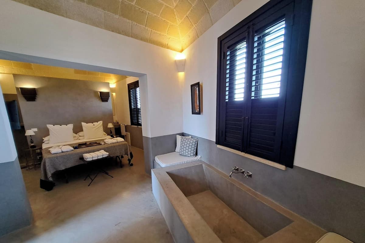 A bedroom with a large bathtub, a bed with white linens, and a window with blinds.