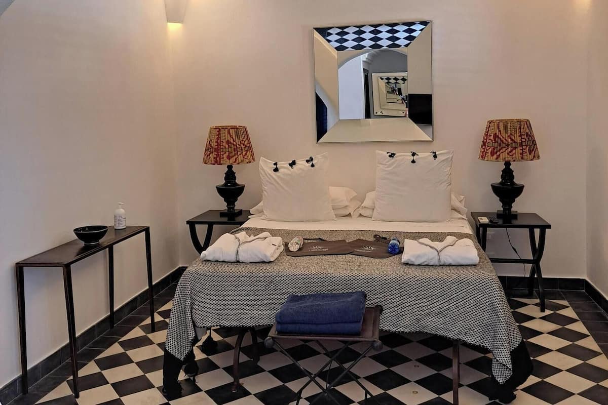 A hotel room with a checkered floor, a bed with white linens, two bedside tables with lamps, and a large mirror on the wall.