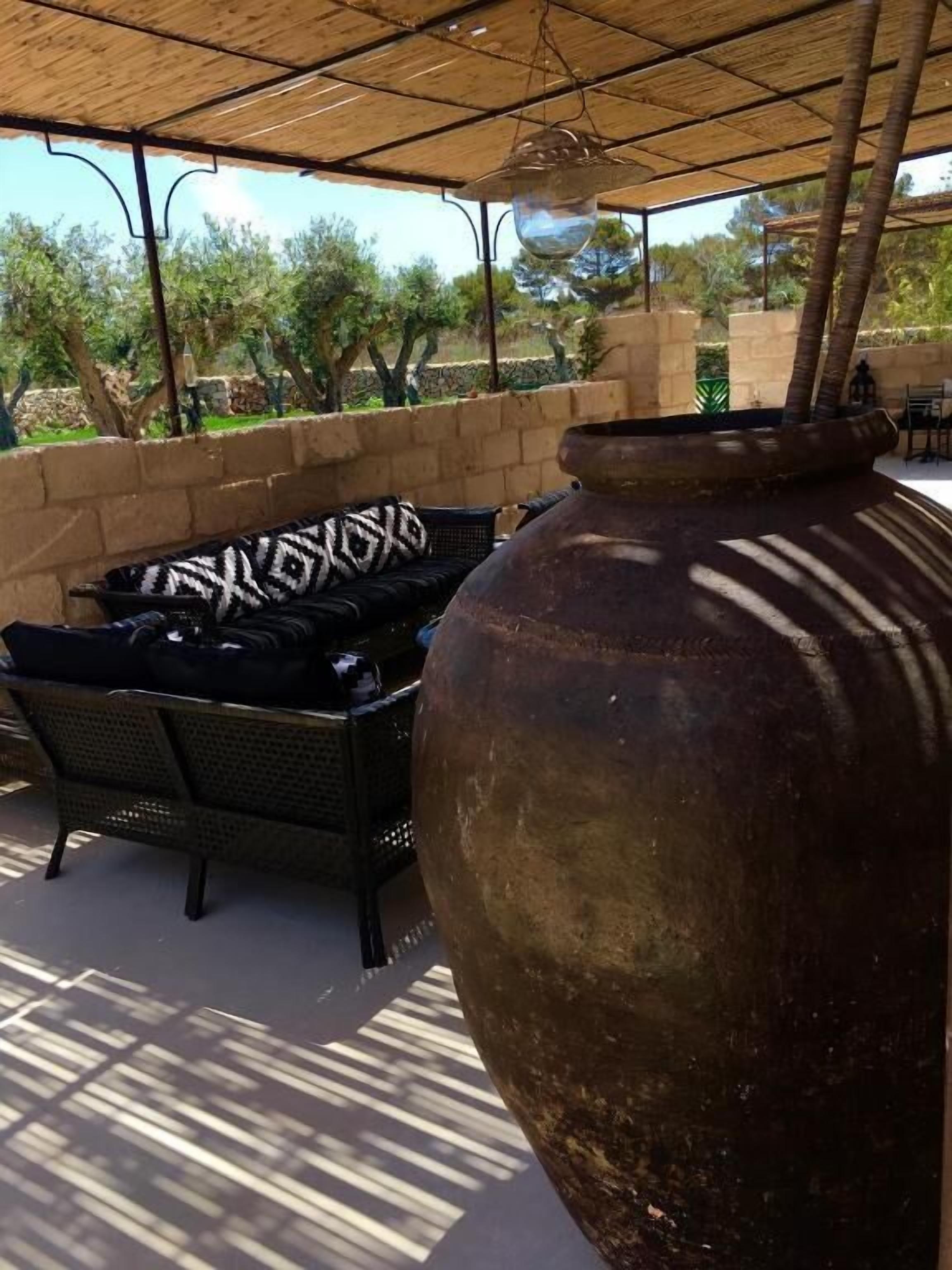 An outdoor patio area with a large ceramic pot, wicker furniture, and a view of trees and a stone wall.