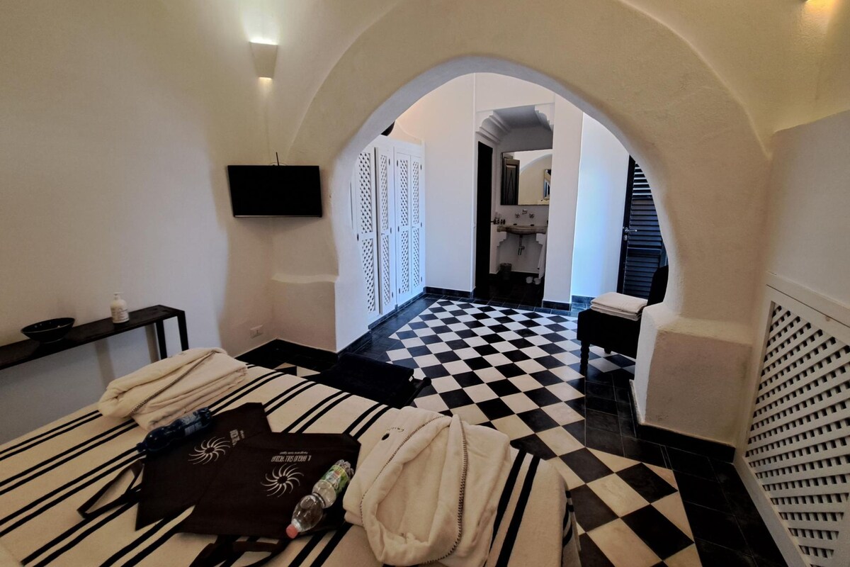 A hotel room with a checkered floor, a TV mounted on the wall, a small table with toiletries, and a bench with towels.