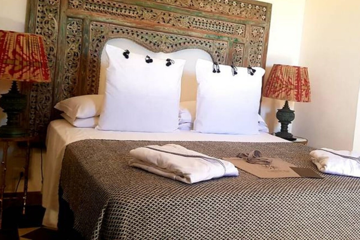 A neatly made bed with white linens, a patterned bedspread, and two decorative pillows. There are two bedside tables with lamps, one on each side of the bed.