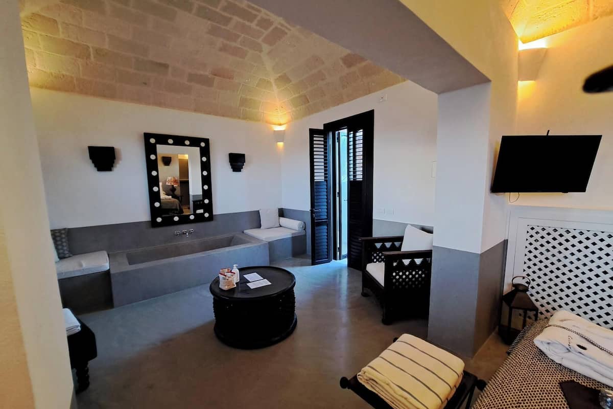 A modern room with a grey sofa, a black round table, a bed with a patterned headboard, a TV, and a mirror with lights.