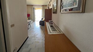 Un pasillo con suelo de baldosas que lleva a una habitación con cama y mesa. Hay cuadros en la pared y una alfombra en el suelo.
