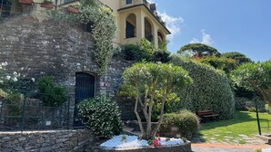 Un edificio de varios pisos con balcones, paredes de piedra y un jardín bien cuidado con diversas plantas y un banco.