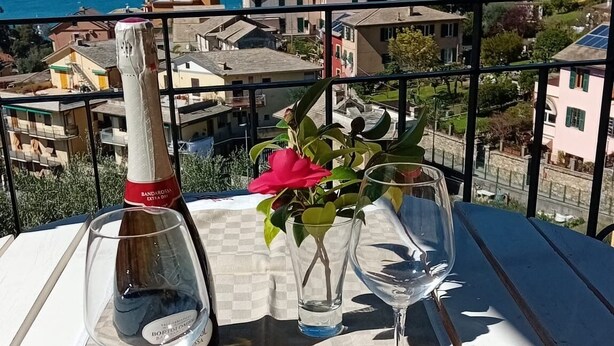 Una mesa con una botella de vino, dos copas y pan, en un balcón con vistas a un pueblo costero.