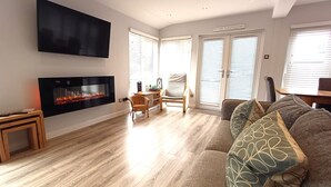 A living room with a fireplace, a flat-screen TV mounted on the wall, a sofa with patterned cushions, and a dining area with a table and chairs.