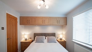A bedroom with a bed, wooden cabinets above, two bedside tables with lamps, a door, and a window with blinds.