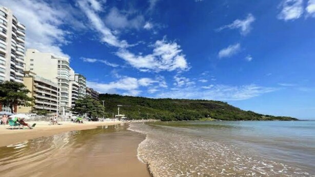 A beach with a sandy shore, a clear blue sky, and a forested hill in the background.