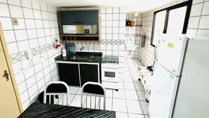 A compact kitchen with white tiled walls, a black countertop, a white sink, a stove, and a refrigerator. There are two white chairs and a small table.