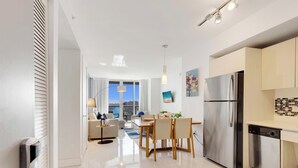 A modern kitchen with a stainless steel refrigerator, a dining area with a table and chairs, and a view of the city through large windows. - Pool + Infinity View w/ Balcony + Gym (Hallandale Beach)