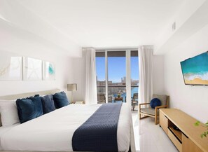 A hotel room with a large bed, a desk, a chair, a TV, and a view of the city and water.