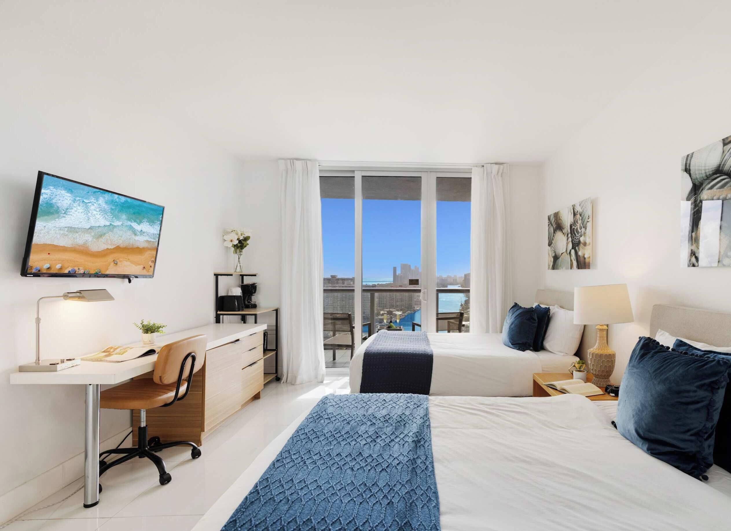 Habitación de hotel con escritorio, dos camas, televisión y vista a la ciudad.