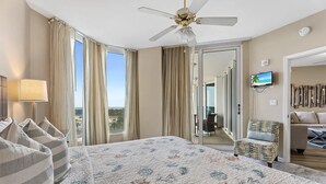A hotel room with a large bed, a ceiling fan, a TV, and a view of the city through the window.