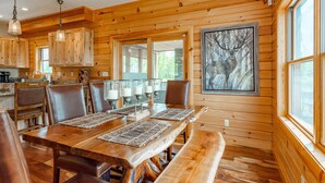 A wooden cabin interior with a dining table, chairs, and a bench. There are wooden cabinets, a painting of a deer, and a window. - Dog Friendly Mountain Lodge w/ Hot Tub (McHenry)