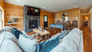 A cozy living room with a stone fireplace, wooden walls, a foosball table, and a large gray sofa. - Dog Friendly Mountain Lodge w/ Hot Tub (McHenry)