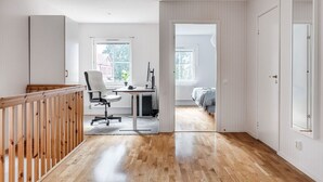 A spacious room with a wooden floor, a desk with a chair, a bed, and a view of a building through a window.