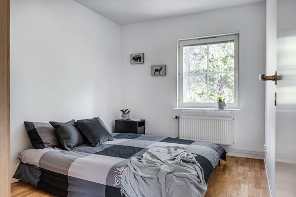 A bedroom with a bed, a window, a radiator, and a small bedside table with a plant.