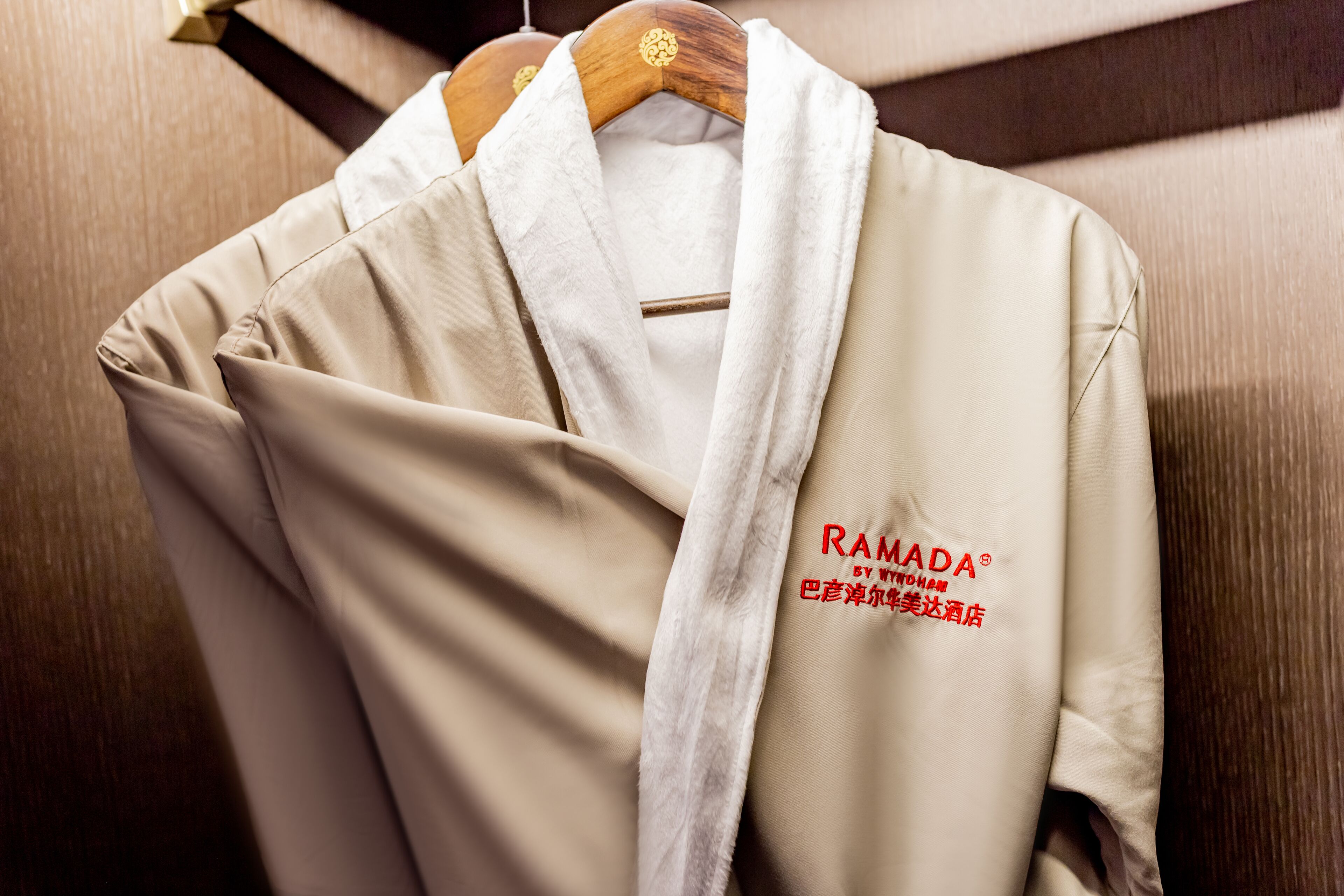 Een beige badjas met de tekst 'RAMADA' en 'BY WU YANG', hangend aan een houten kleerhanger.