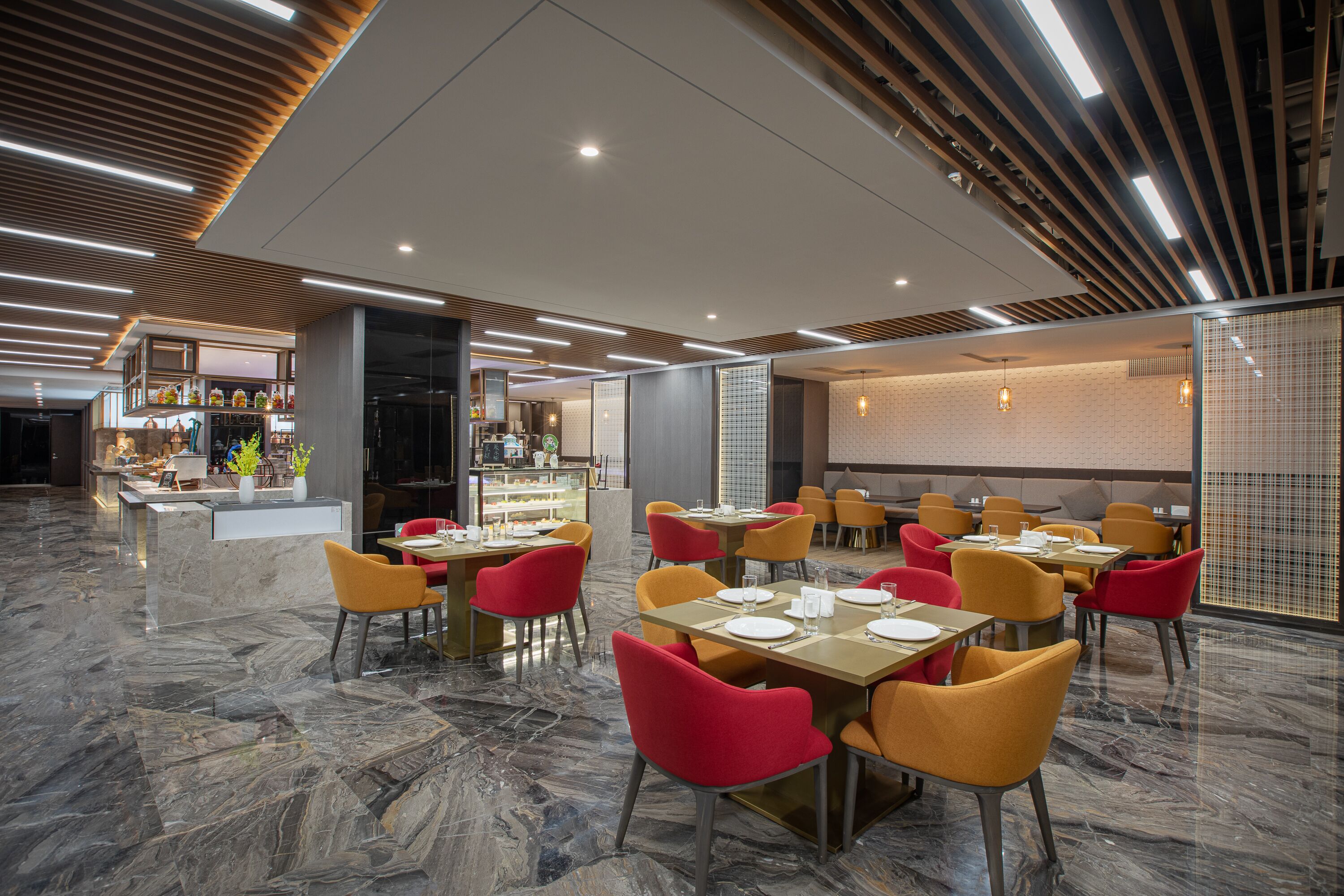 Restaurante moderno com chão de mármore, cadeiras vermelhas e amarelas, e um balcão exibindo diversos alimentos.