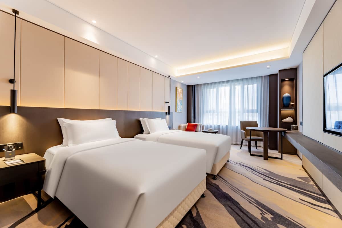 Habitación de hotel con dos camas, un escritorio, una silla y un amplio ventanal con cortinas.