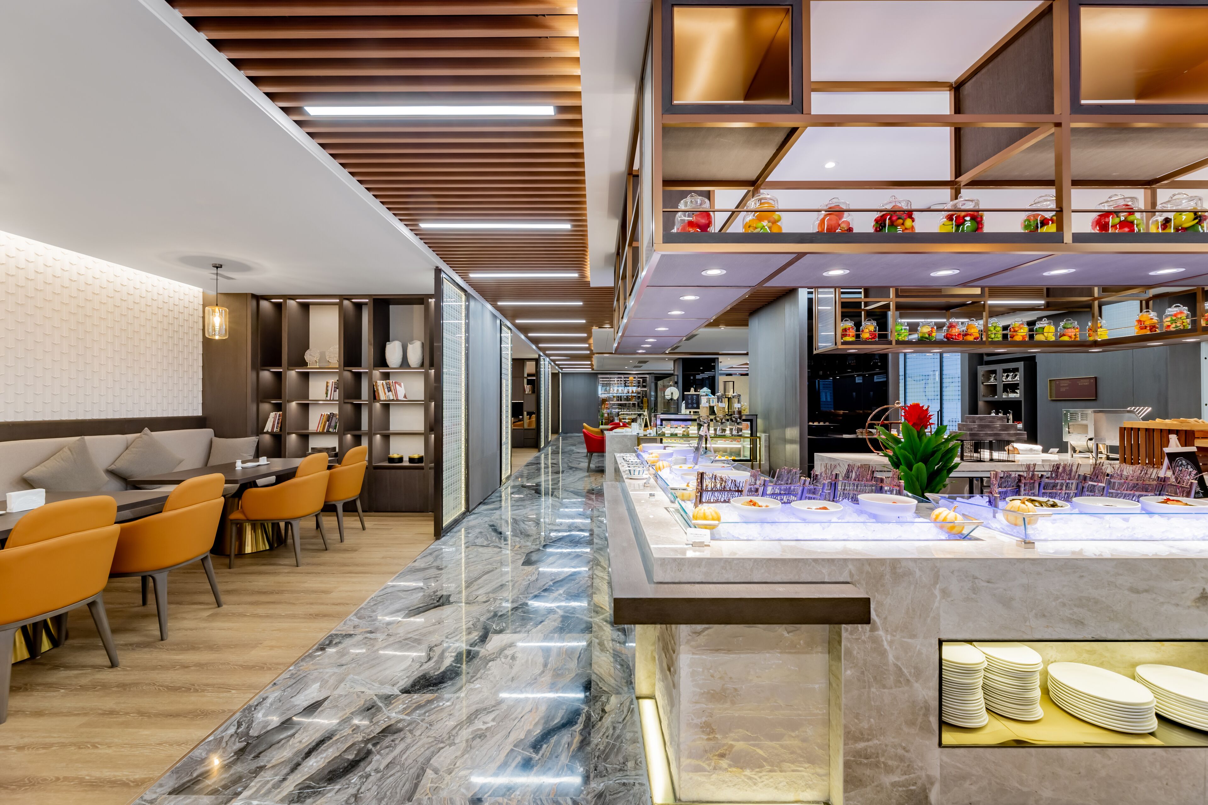 A modern hotel lobby with a marble reception desk, seating area with orange chairs, and a buffet counter with various food items.